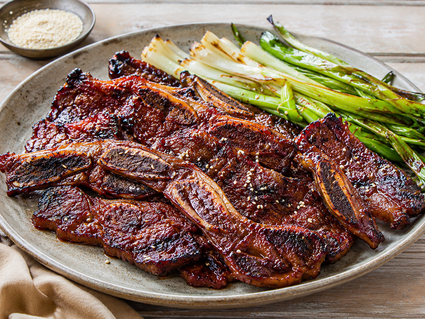 Glazed bone-in beef short ribs with charred edges, sticky dark barbecue glaze, sesame seeds and scallions on a white plate