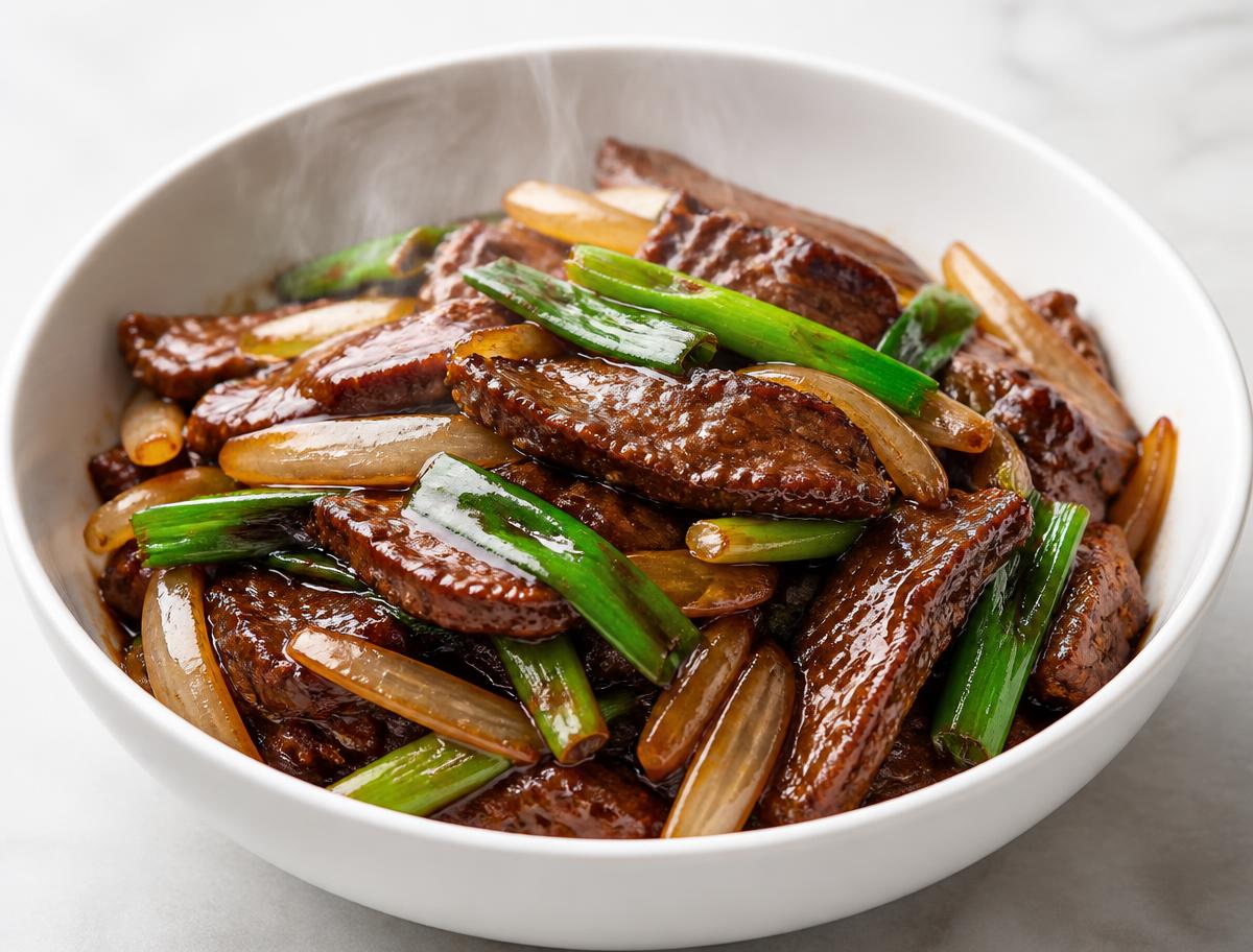 Mongolian beef with tender sliced steak, onions and green onions in a glossy dark savory glaze in a white bowl